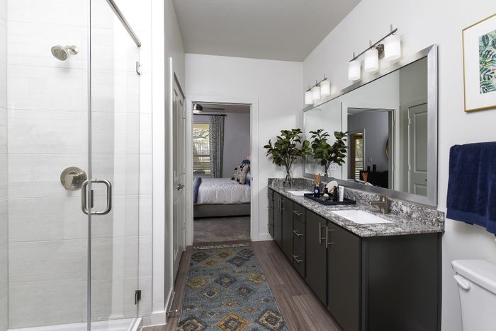 Stylish bathroom featuring a double sink vanity with elegant granite countertops and a walk-in shower, leading to a cozy bedroom with modern decor.