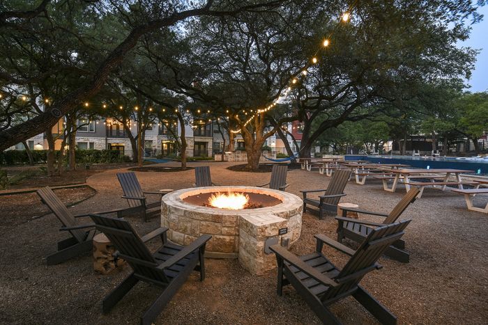 Charming outdoor space with a cozy stone fire pit, surrounded by Adirondack chairs, twinkling string lights, and a backdrop of lush trees, perfect for evening gatherings.