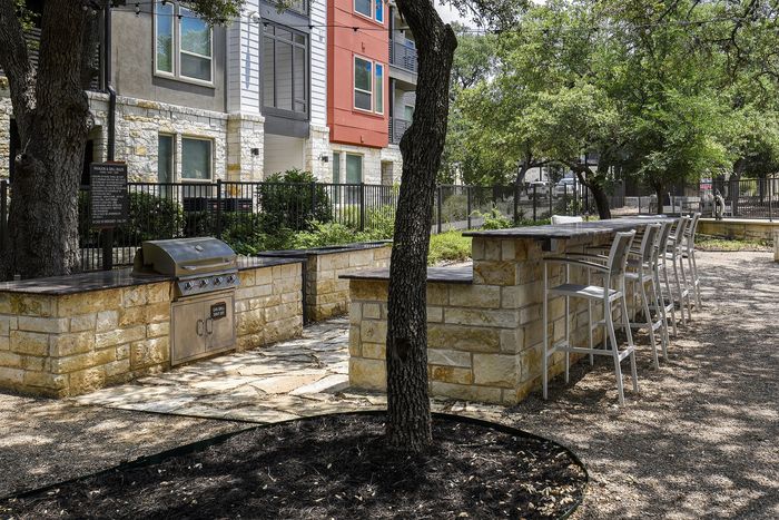 Inviting outdoor space featuring a stone BBQ grill area with modern bar seating, surrounded by lush greenery and shaded by mature trees.