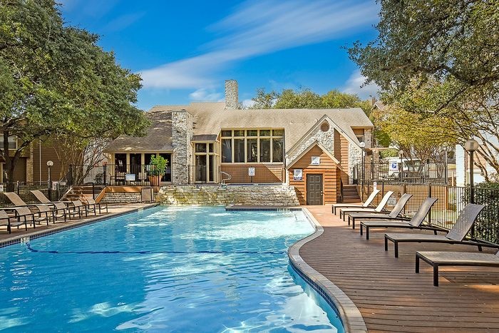 Inviting apartment community pool with sun loungers, surrounded by lush trees and a cozy clubhouse under a clear blue sky.