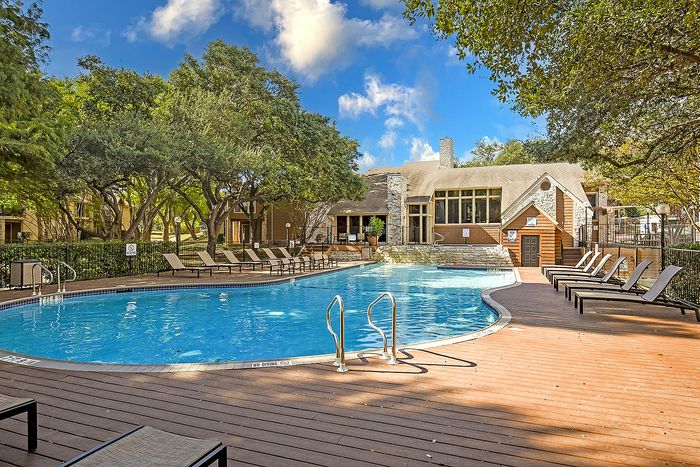 Luxurious apartment complex featuring a sparkling swimming pool, surrounded by lush trees and stylish lounge chairs under a vibrant blue sky.
