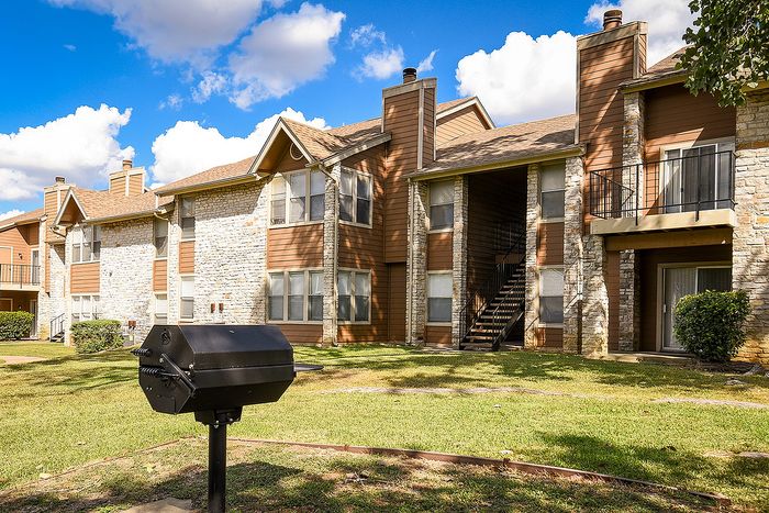 Charming apartment complex with rustic stone facade, featuring lush green lawns and barbecue area under a bright blue sky.