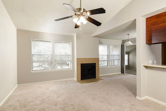 Inviting living room with plush carpeting, large windows, stylish ceiling fan, and a cozy fireplace, perfect for a comfortable home atmosphere.