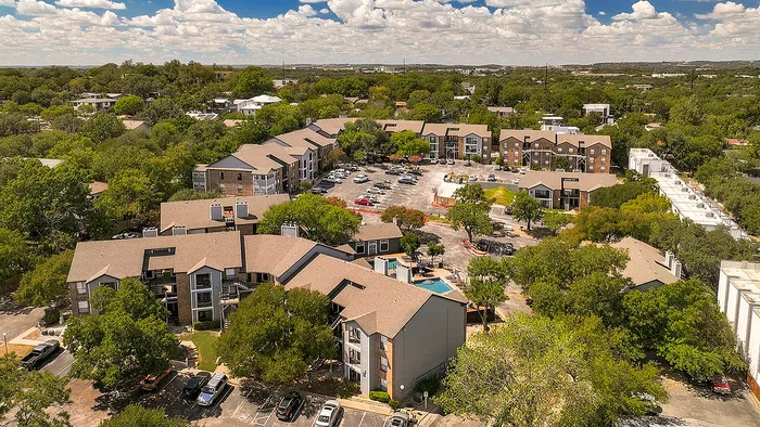 Charming aerial view of a picturesque apartment complex surrounded by lush greenery, featuring multiple modern buildings, ample parking, and serene outdoor spaces.