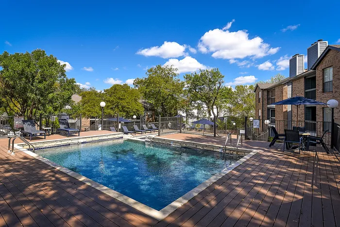 Charming apartment community featuring a sunlit swimming pool with a spacious deck and cozy outdoor seating, surrounded by lush greenery.