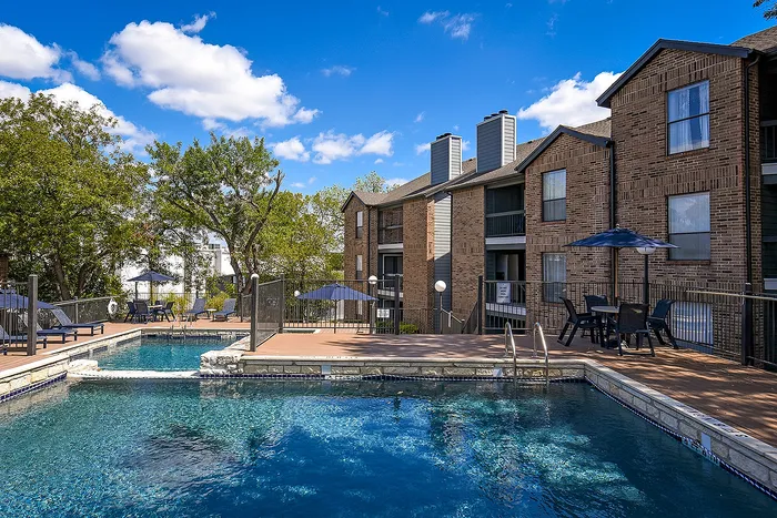 Inviting apartment complex with a refreshing outdoor pool, surrounded by lush trees and modern patio seating under a clear blue sky.