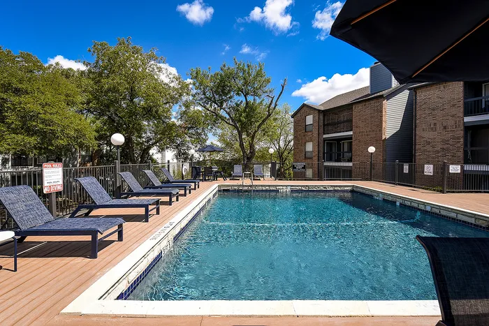 Inviting outdoor pool area with modern lounge chairs, surrounded by lush greenery and elegant apartment buildings under a bright blue sky.