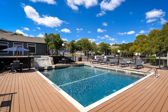 Inviting outdoor swimming pool area with sun loungers, wooden deck, and vibrant landscaping under a bright blue sky.