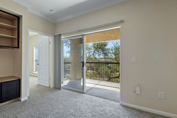 Bright apartment interior featuring plush carpeting, built-in shelving, and a sliding glass door opening to a scenic balcony overlooking lush greenery.