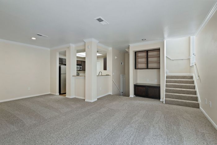 Bright and open apartment living space featuring plush carpet, modern kitchen with stainless steel appliances, and elegant built-in shelving beside a stairway.