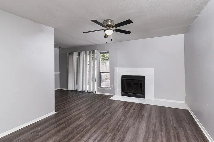 Modern living room with sleek wood flooring, a cozy fireplace, and elegant ceiling fan, enhanced by natural light from floor-to-ceiling windows with stylish blinds.