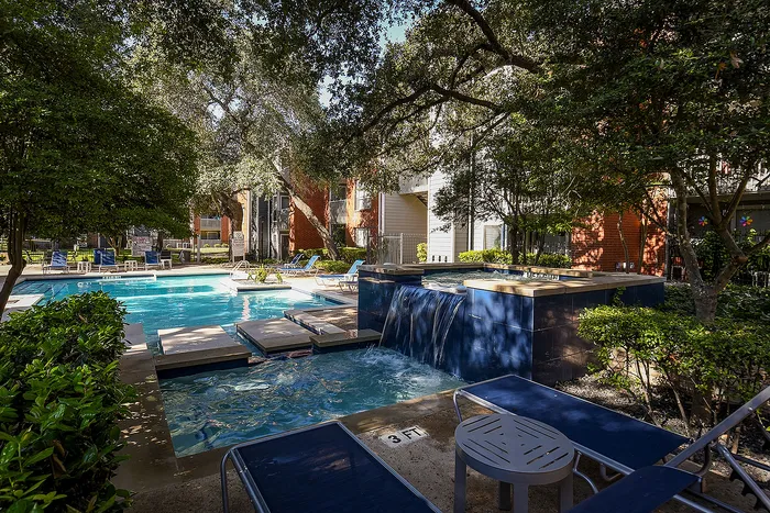 Serene apartment complex pool area with lush greenery, a cascading water feature, and ample lounge seating, ideal for relaxation and leisure.
