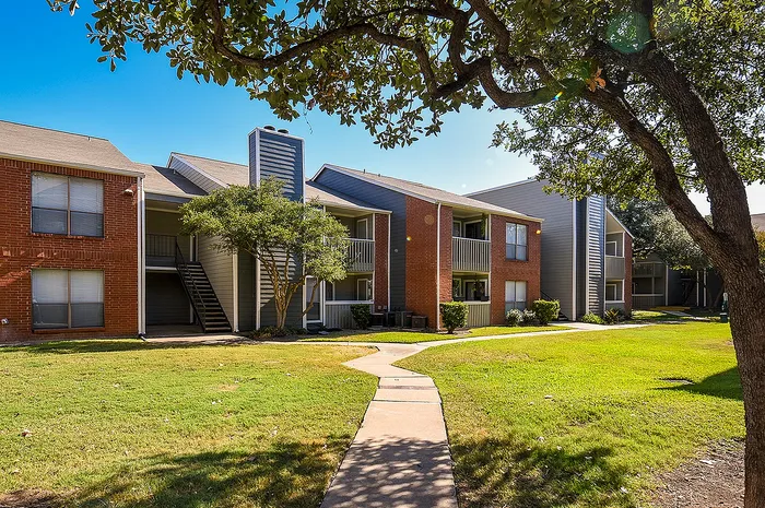 Charming apartment complex with brick exteriors, lush green lawns, and mature trees under a clear blue sky.