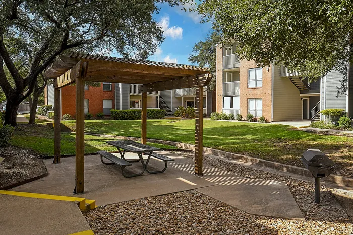 Charming apartment complex courtyard featuring a shaded pergola with picnic table, lush green lawn, and beautifully maintained buildings.