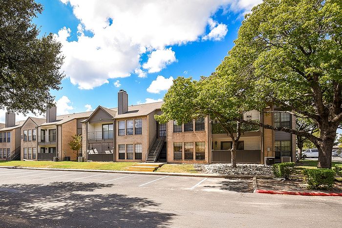 Charming apartment complex surrounded by lush trees and bright blue skies, featuring spacious balconies and ample parking.
