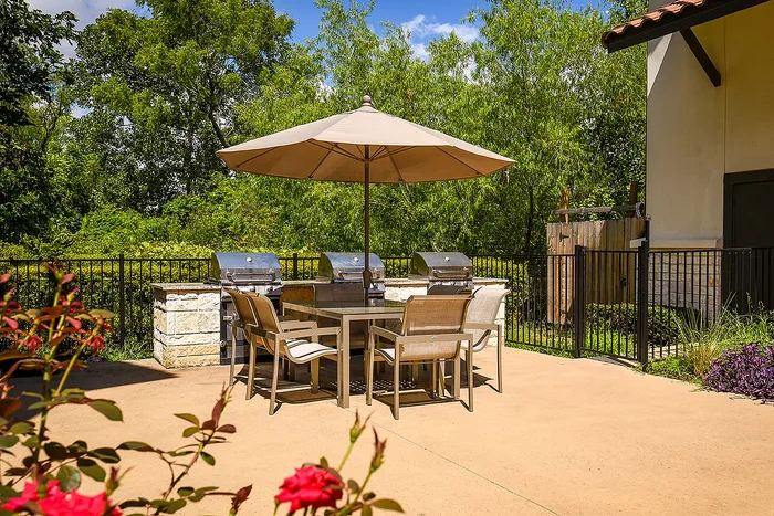 Inviting outdoor patio with a modern table, chairs, and stainless steel grills under a large umbrella, surrounded by lush greenery.