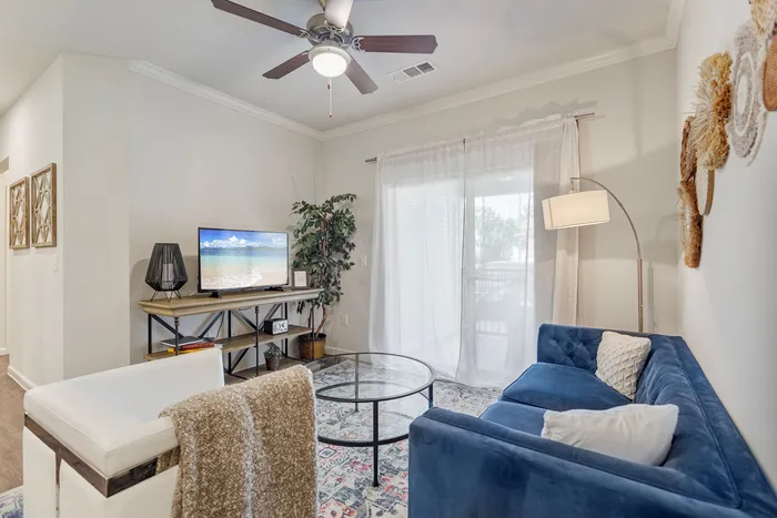 Bright and inviting living room featuring a blue velvet sofa, modern glass coffee table, and a stylish entertainment area with a flat-screen TV and lush greenery.