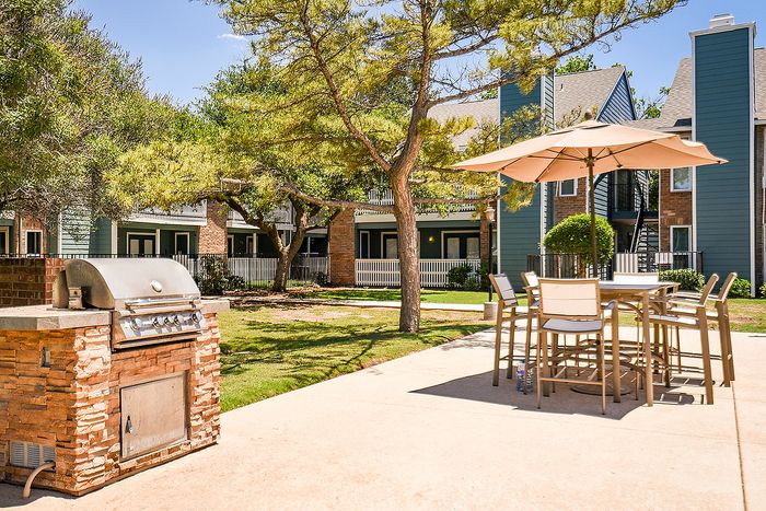 Charming outdoor patio with a modern grill, shaded dining area, and lush greenery at an apartment complex.