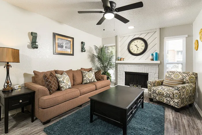 Inviting living room with cozy brown sofa, decorative pillows, elegant armchair, and a modern coffee table. Features a stylish ceiling fan, framed art, and a charming fireplace with a large clock, creating a perfect space for relaxation.
