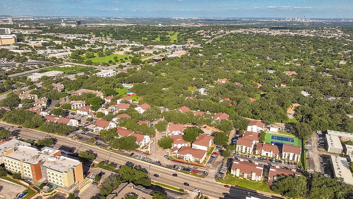 Expansive aerial view showcasing a vibrant residential neighborhood surrounded by lush greenery, with modern apartment complexes and a tennis court at the heart of the community.