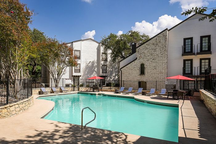 Elegant apartment complex featuring a sunlit swimming pool surrounded by comfortable lounge chairs and vibrant red umbrellas.