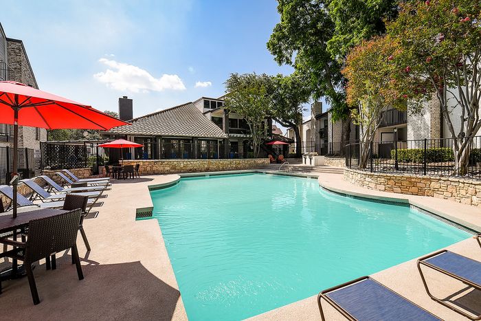 Stunning apartment community pool area with crystal-clear water, stylish lounge chairs, and vibrant red umbrellas, offering a luxurious outdoor retreat.