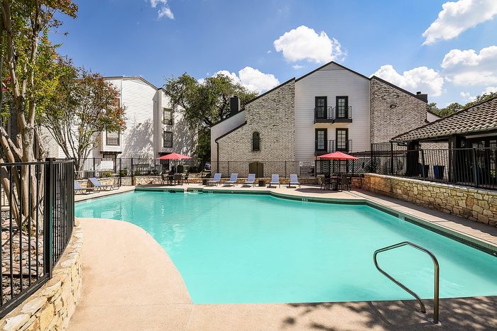 Elegant apartment complex featuring a clear blue swimming pool, relaxing loungers, and charming stone architecture under a bright, sunny sky.