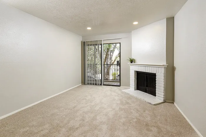 Inviting living room with plush carpeting, modern recessed lighting, and a charming white brick fireplace next to sliding glass doors leading to a serene outdoor view.