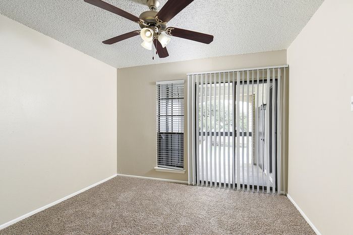 Bright and airy room with plush carpet, ceiling fan, and large windows leading to a balcony, perfect for relaxing.