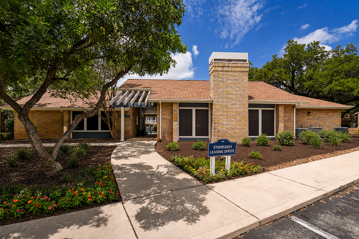 Inviting leasing office surrounded by lush greenery and vibrant flowers, featuring a charming brick exterior and shaded pathway.