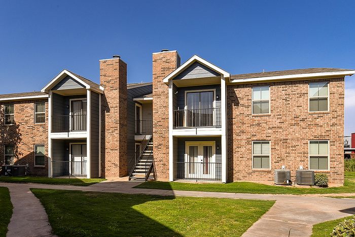 Charming brick apartment building with modern balconies, surrounded by lush green lawns and clear blue skies.