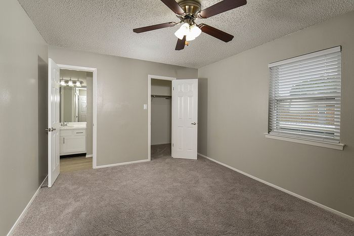 Inviting bedroom with plush carpet, large window with blinds, and ceiling fan; featuring a closet and connecting door to a sleek bathroom with modern vanity.