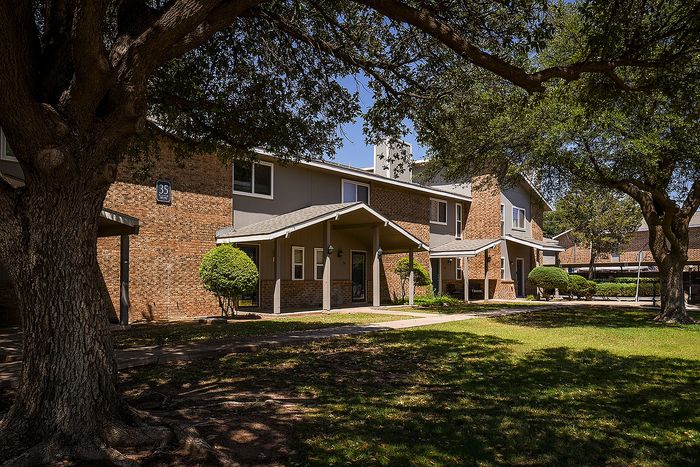Charming two-story brick apartment complex nestled among lush greenery and mature trees, offering a serene and inviting atmosphere.