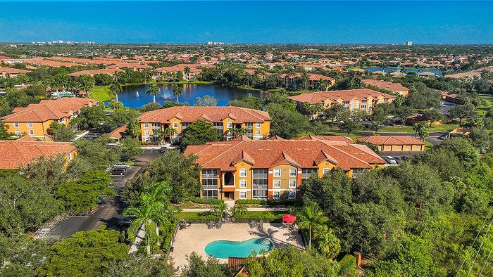Aerial view of a picturesque apartment complex with terracotta roofs, lush greenery, a sparkling pool, and serene lake with fountain.