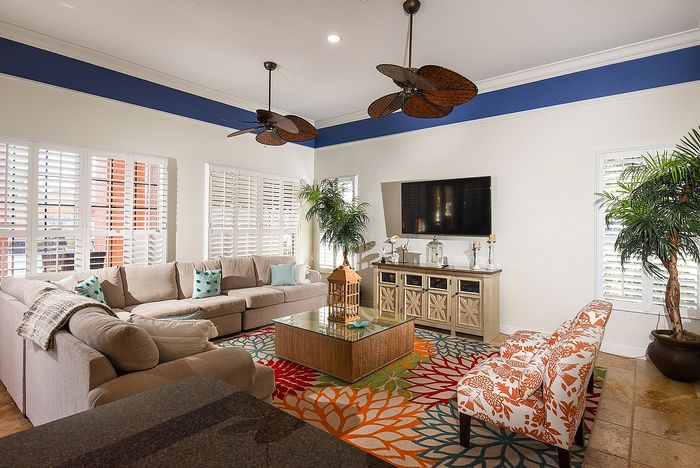 Inviting living room with plush sectional sofa, colorful floral rug, and large TV, illuminated by natural light and accentuated with plantation shutters.