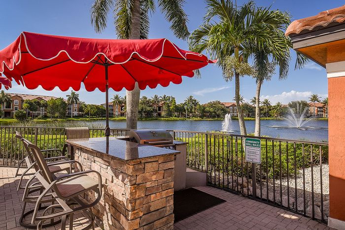 Charming outdoor patio with red umbrella, overlooking a tranquil lake and fountains, surrounded by lush palm trees and serene apartment views.