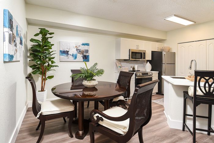 Elegant dining area with a round wooden table and stylish chairs, adjacent to a modern kitchen equipped with stainless steel appliances and chic white cabinetry. Bright artwork and lush greenery enhance the inviting atmosphere.