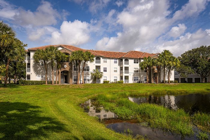 Charming apartment building with lush palm trees and serene water view under a bright blue sky.