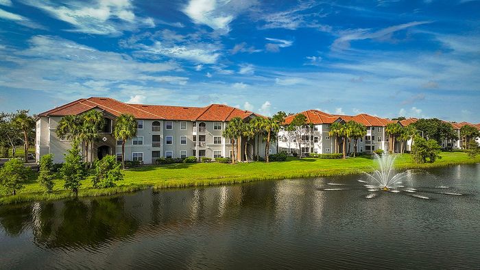 Charming apartment buildings with red-tiled roofs nestled beside a tranquil pond and fountain, surrounded by lush greenery and palm trees under a vibrant blue sky.