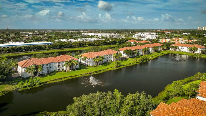 Scenic aerial view of apartment complex with red-tile roofs, surrounded by lush greenery and a serene lake with fountains.