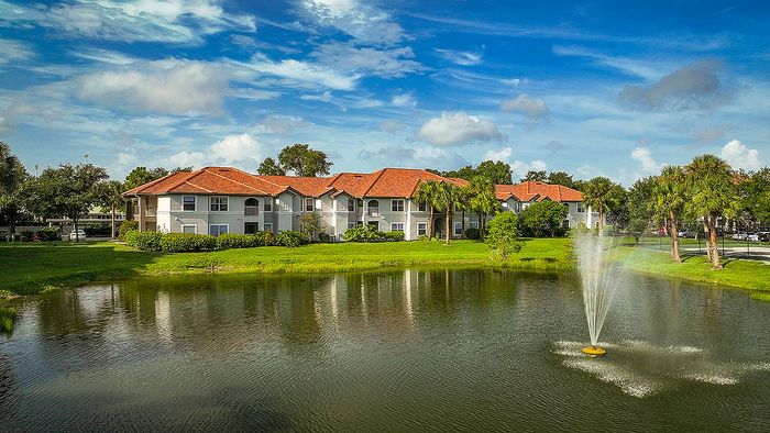Charming lakeside apartment complex with red tile roofs, lush greenery, and a fountain under a vibrant blue sky.