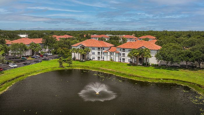 Charming apartment complex with red-tiled roofs overlooking a serene pond with a central fountain and lush greenery, offering a tranquil living environment.