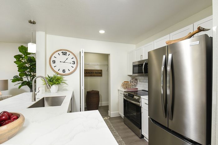Modern kitchen with sleek stainless steel appliances, elegant white cabinetry, and a stylish open layout. Features a large wall clock, tasteful plants, and convenient access to a dedicated laundry room.