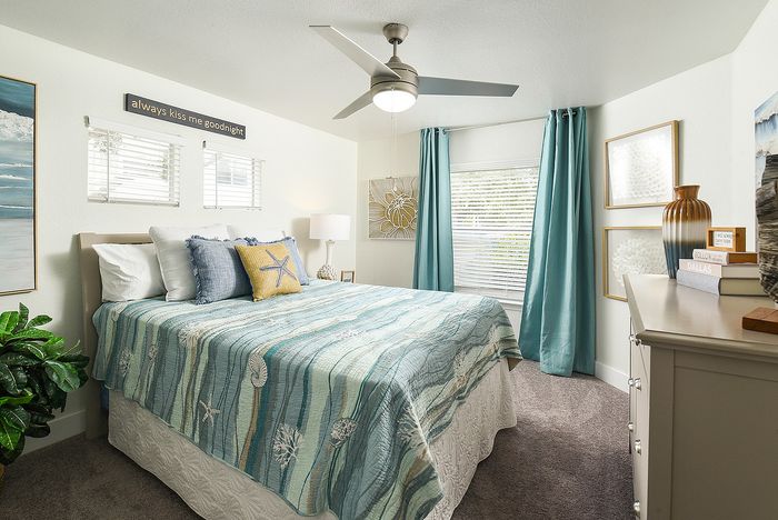 Charming bedroom with coastal decor, featuring a stylish blue comforter, modern ceiling fan, and sunlit windows with elegant teal curtains.
