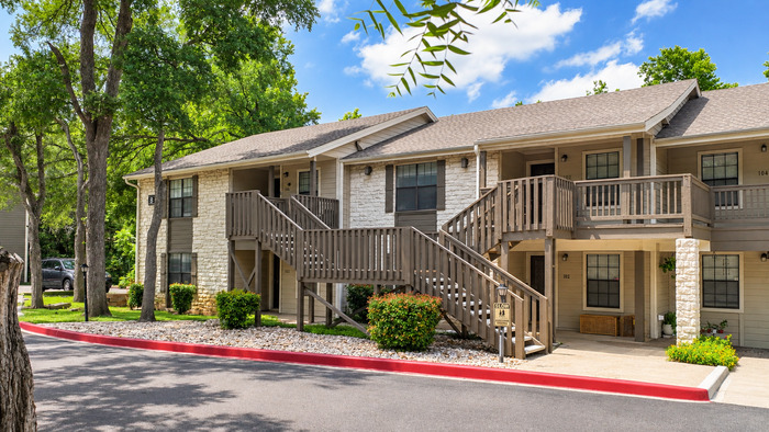 Charming two-story apartment complex with stone and wood exterior, surrounded by lush greenery and shaded by tall trees on a sunny day.