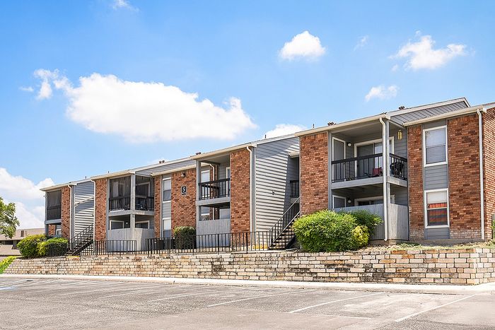 Inviting apartment complex with modern brick design, lush greenery, and ample parking under a vibrant blue sky.