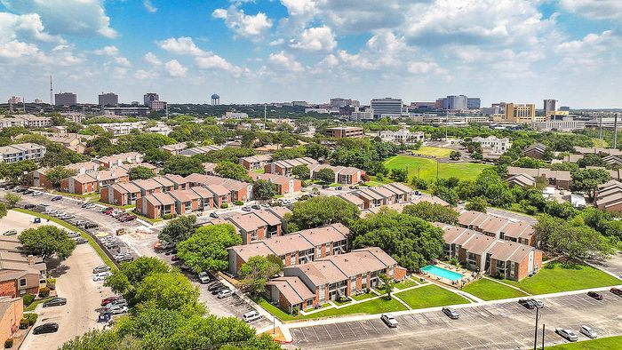 Vibrant aerial view of a charming residential community with contemporary apartments, lush greenery, and a sparkling swimming pool, set against a backdrop of cityscape.