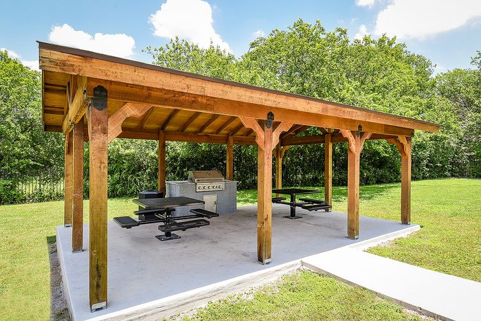 Inviting outdoor pavilion with a wooden roof, featuring a modern grill and picnic-style seating, surrounded by lush greenery.