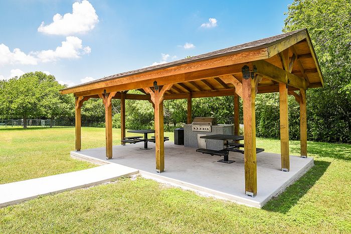 Charming wooden pavilion with picnic tables and a grill, set in a lush green park, perfect for outdoor gatherings.