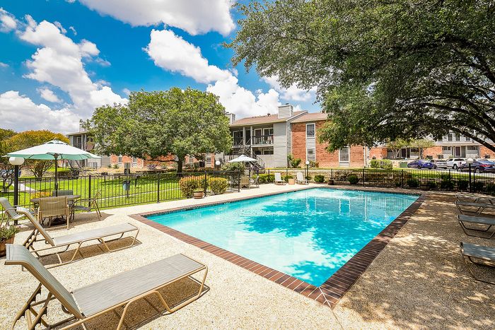 Inviting apartment complex pool area with lounge chairs and umbrellas, surrounded by lush greenery and beautiful trees under a vibrant blue sky.